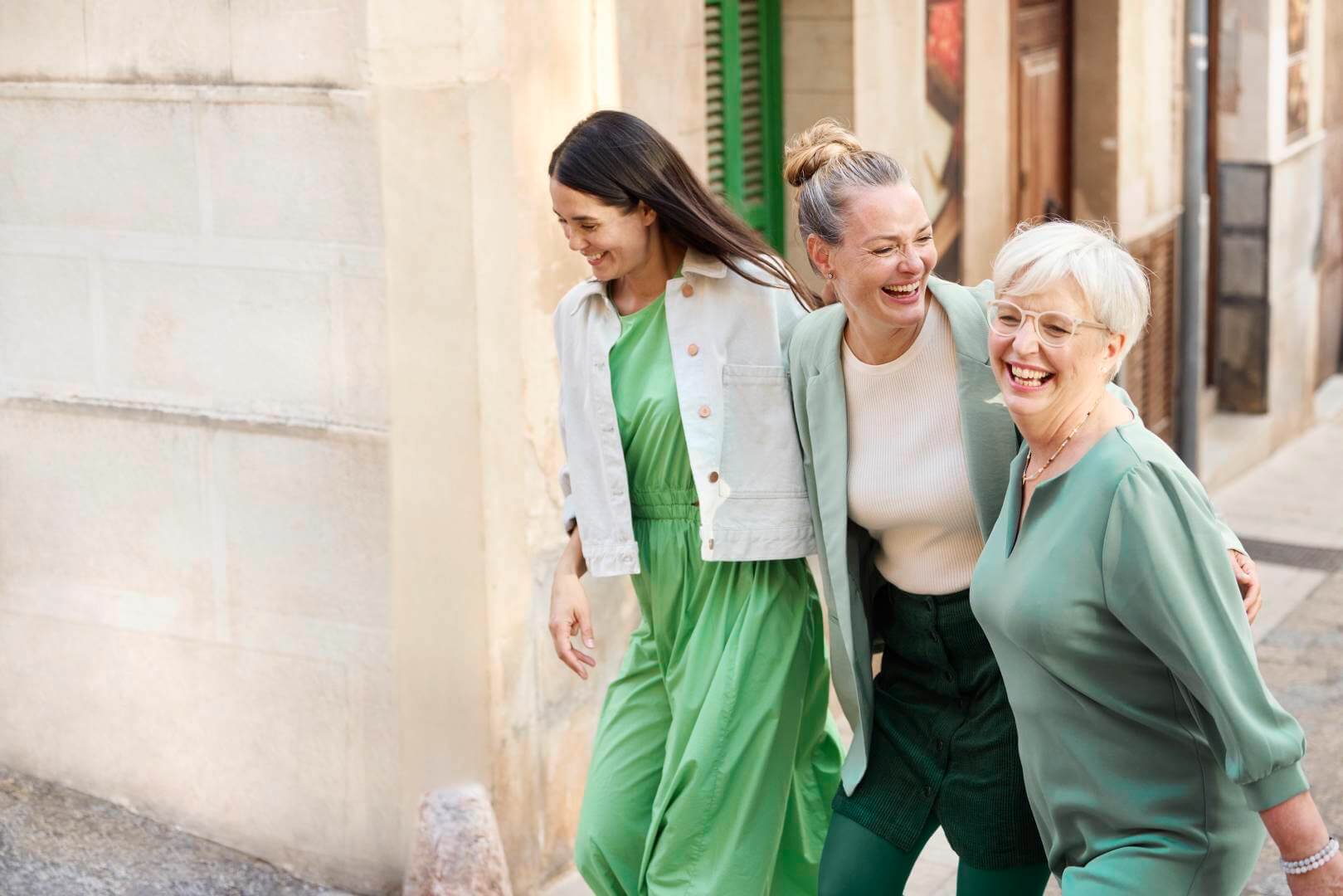 Drei Frauen unterschiedlicher Generationen lachen gemeinsam, während sie Arm in Arm durch eine mediterrane Altstadt gehen. Sie tragen stilvolle Outfits in verschiedenen Grüntönen, die harmonisch aufeinander abgestimmt sind.